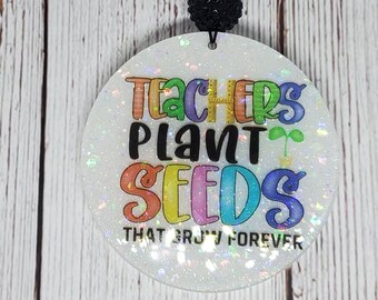 May include: A round, glittery white car charm with the text "TEACHERS PLANT SEEDS THAT GROW FOREVER" in colourful letters. The charm is attached to a black string with a sunflower bead and a black beaded accent.