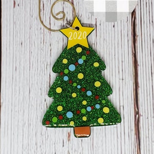 May include: A Christmas tree ornament in the shape of a green glittery tree with colorful dots and a yellow star on top with the year 2020. It has a gold-colored hook for hanging.