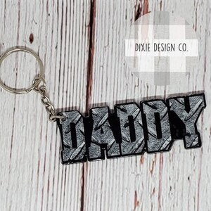 May include: A silver keychain with the word "DADDY" in black and gray diamond plate design. The keychain has a silver ring and chain. The background is a light wood grain.