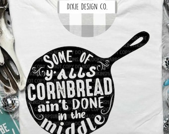 May include: White t-shirt with a black graphic of a cast iron skillet with the text "Some of y'alls cornbread ain't done in the middle". The text is in a vintage font style. The graphic is surrounded by a decorative border.