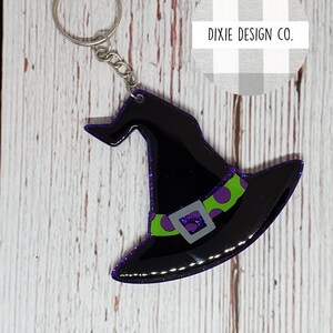 May include: A black witch hat keychain with a purple glitter trim, green band with purple polka dots, and a silver buckle. The keychain has a silver ring and chain. The text "DIXIE DESIGN CO." is in the background.