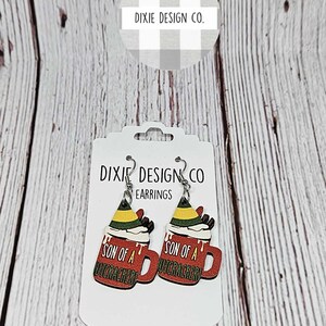 May include: A pair of wooden earrings featuring a red mug with white whipped cream and a green elf hat. The mug says "Son of a Nutcracker".