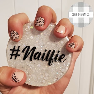 May include: A round, glittery white coaster with the hashtag #Nailfie in black script. The nails in the image are painted with a leopard print design and a solid nude color. The Dixie Design Co. logo is in the background.
