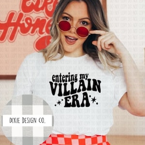 May include: White t-shirt with the text "entering my VILLAIN ERA" in black, with two star graphics. The shirt is worn with a red and white checkered skirt. The image also includes a logo for Dixie Design Co.