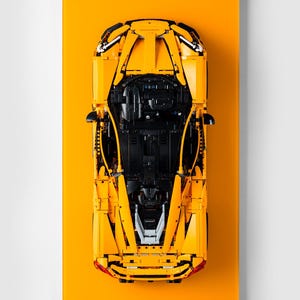 May include: A top-down view of a bright yellow sports car model, set against a matching yellow background. The car's intricate details, including black interior components and red taillights, are clearly visible. The model is a detailed replica of a sports car.