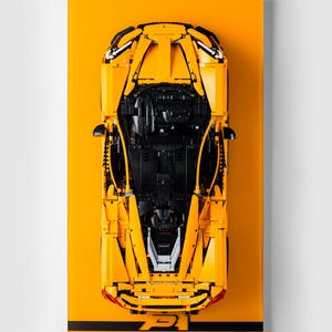 Peut inclure: Vue de dessus d'une voiture modèle Lego jaune vif, sur un fond jaune assorti. La voiture présente des détails noirs et est montée sur une base rectangulaire avec une bande noire en bas. Le modèle est une réplique détaillée.