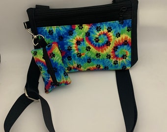 Puede incluir: Un bolso bandolera negro con un estampado tie-dye con huellas de patas. El bolso tiene una cremallera y una bolsa extraíble con una cremallera.