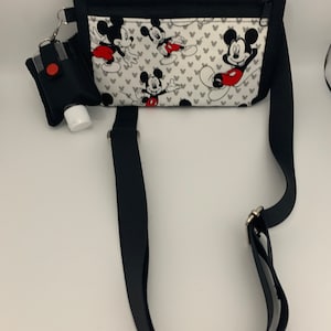 May include: Black crossbody bag with a white fabric insert featuring a Mickey Mouse print. The bag has a zipper closure and a black strap. A small black pouch with a red button is attached to the bag.