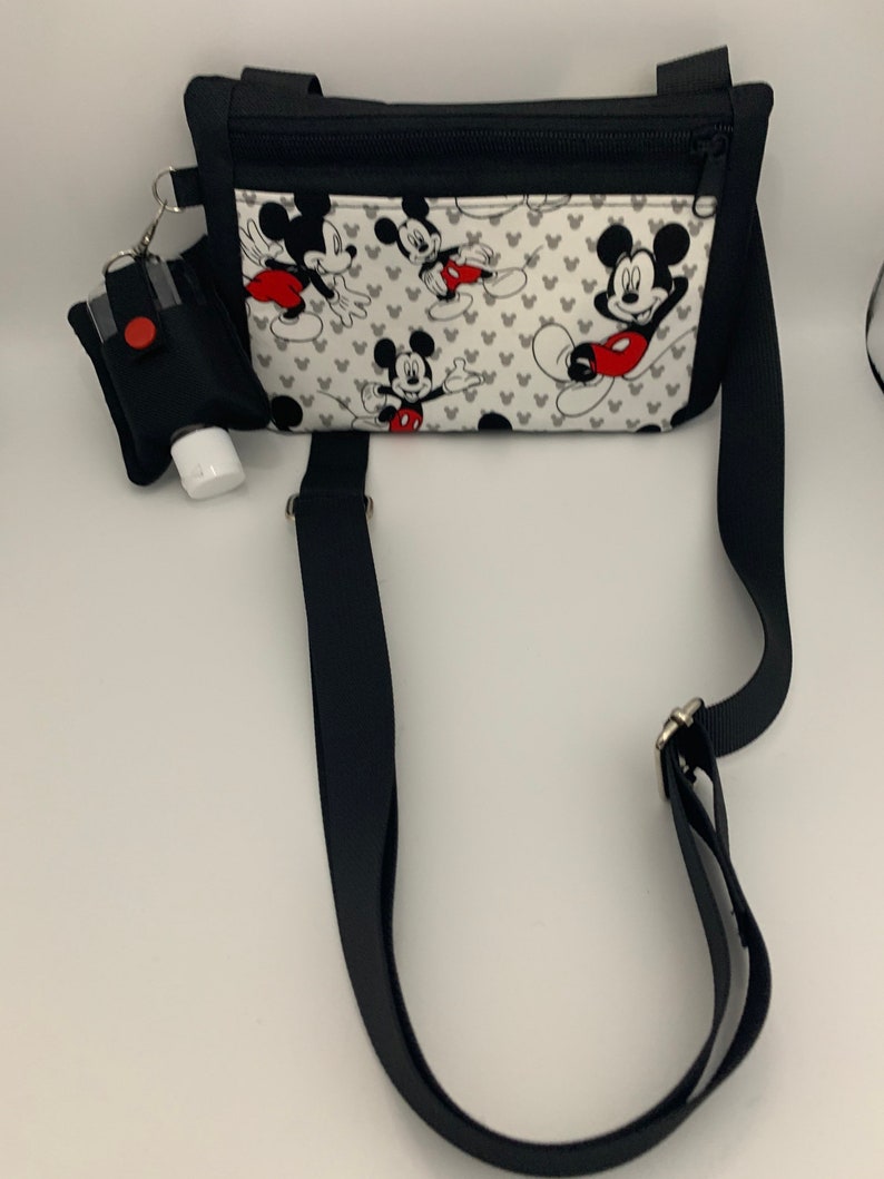 May include: Black crossbody bag with a white fabric insert featuring a Mickey Mouse print. The bag has a zipper closure and a black strap. A small black pouch with a red button is attached to the bag.