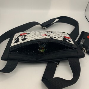 May include: A black crossbody bag with a white fabric lining featuring a Mickey Mouse and Minnie Mouse print. The bag has a zipper closure and a black adjustable strap.