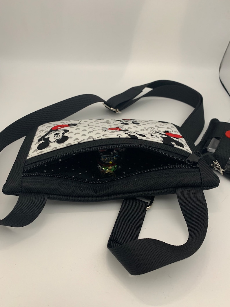 May include: A black crossbody bag with a white fabric lining featuring a Mickey Mouse and Minnie Mouse print. The bag has a zipper closure and a black adjustable strap.