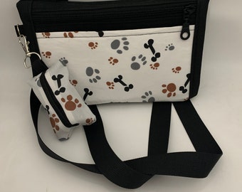 Puede incluir: Un bolso bandolera blanco y negro con un diseño de huellas de patas y huesos. El bolso tiene cierre de cremallera y un pequeño bolsillo adjunto.
