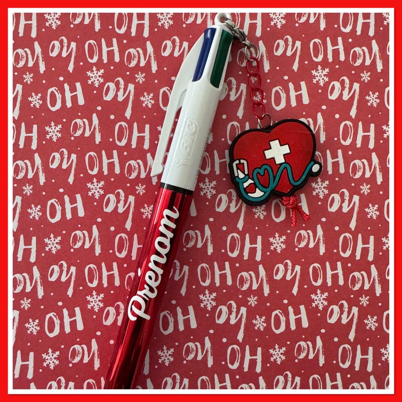 Puede incluir: Un bol&iacute;grafo rojo y blanco con la palabra "Prenom" en escritura blanca. El bol&iacute;grafo tiene un dije con un coraz&oacute;n rojo, una cruz blanca y un estetoscopio. El fondo es rojo con copos de nieve blancos y las palabras "OH" y "HO".