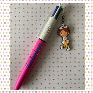 Puede incluir: Un bolígrafo BIC rosa con el nombre "Sabrina" en escritura azul. El bolígrafo tiene un cuerpo blanco y una punta multicolor. Un charm de una enfermera de dibujos animados está unido al bolígrafo con una pequeña cadena. El fondo es una superficie blanca con patrones de corazones.