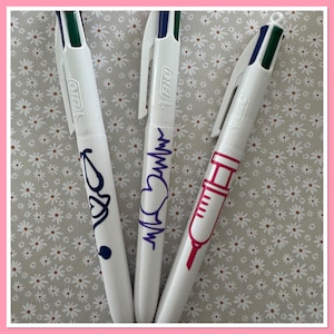 Puede incluir: Tres bolígrafos retráctiles blancos con diferentes diseños de temática médica. Los bolígrafos tienen tinta azul, verde y negra. Un bolígrafo tiene un estetoscopio y un corazón, otro tiene una línea de latido del corazón y un corazón, y el último tiene una jeringa.