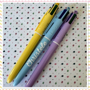 Puede incluir: Tres bolígrafos multicolores con el nombre de la marca "BIC" impreso en ellos. Los bolígrafos son amarillos, azul claro y lavanda. El bolígrafo azul claro tiene el nombre "Christèle" escrito en él.