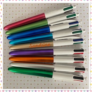 Puede incluir: Un conjunto de diez bolígrafos multicolores con cuerpos blancos y tinta negra, roja, azul y verde. Los bolígrafos tienen acabados metálicos en colores como dorado, plateado, morado, naranja, verde, azul y rosa. Los bolígrafos están dispuestos verticalmente con el cuerpo de cada bolígrafo mirando hacia la cámara. El nombre "Sandrine" está escrito en uno de los bolígrafos.