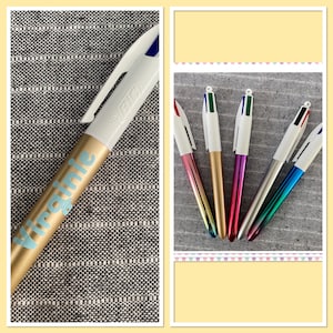Puede incluir: Una colección de bolígrafos BIC con barriles blancos y partes inferiores metálicas coloridas. Un bolígrafo es dorado con el nombre "Virginie" en azul claro. Los otros bolígrafos tienen puntas rojas, verdes, azules y negras.