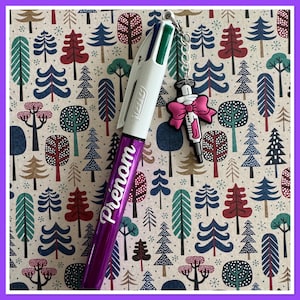 Pode incluir: Uma caneta multicolorida com a palavra "Pr&eacute;nom" em escrita branca em um corpo roxo. Um pingente de seringa com um la&ccedil;o rosa pende da caneta. O fundo apresenta um padr&atilde;o de &aacute;rvores estilizadas.