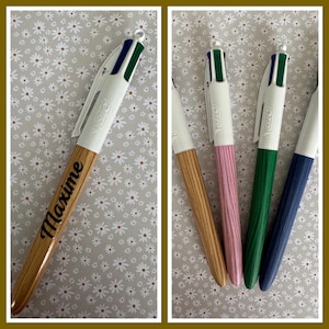 Puede incluir: Cuatro bolígrafos multicolores con barriles de madera. Los bolígrafos son de diferentes colores: madera natural, rosa, verde y azul. El bolígrafo de la izquierda tiene el nombre "Maxime" grabado en el barril.
