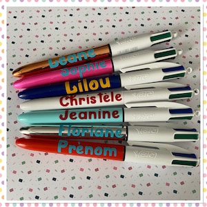 Puede incluir: Un conjunto de diez bolígrafos multicolores con diferentes nombres escritos en ellos. Los bolígrafos son todos blancos con tinta azul, verde y negra. Los nombres en los bolígrafos son: Leane, Sophie, Lilou, Christelle, Jeanine, Floriane y Prenom.