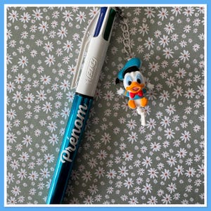 Peut inclure: Un stylo multicolore BIC bleu et blanc avec le mot "Pr&eacute;nom" imprim&eacute; sur le corps. Un charm Donald Duck est attach&eacute; au stylo avec une cha&icirc;ne argent&eacute;e. Le fond est un motif floral.