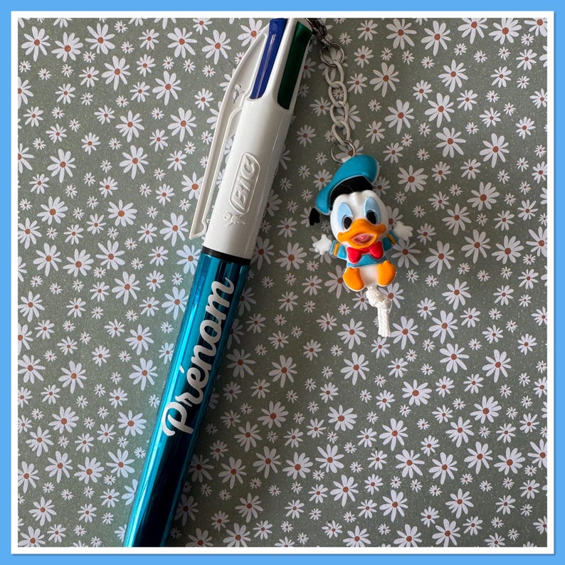 Peut inclure: Un stylo multicolore BIC bleu et blanc avec le mot "Pr&eacute;nom" imprim&eacute; sur le corps. Un charm Donald Duck est attach&eacute; au stylo avec une cha&icirc;ne argent&eacute;e. Le fond est un motif floral.