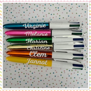 Puede incluir: Seis bolígrafos personalizados con cuerpos de diferentes colores. Los bolígrafos son azules, rosas, verdes, dorados, naranjas y amarillos. Cada bolígrafo tiene un cuerpo blanco con una inscripción negra. Las inscripciones dicen: Virginie, Melanie, Florian, Christine, Clem y Jannot.