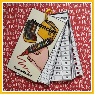 May include: A color swatch book with a yellow ring, featuring illustrations of a paint palette, a suitcase, and an acrylic marker. The text "Nuancier" and "GuangNa 360" are visible. The background is a festive red and white pattern.