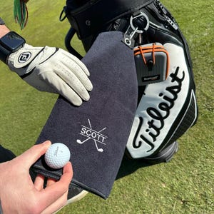 May include: A black golf towel with white embroidery that says "SCOTT" and has a pair of crossed golf clubs. The towel is hanging on a black and white golf bag.