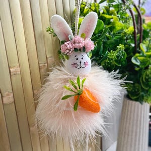 May include: A pink fluffy Easter bunny with a flower crown and a carrot in its hand. The bunny is hanging from a string.