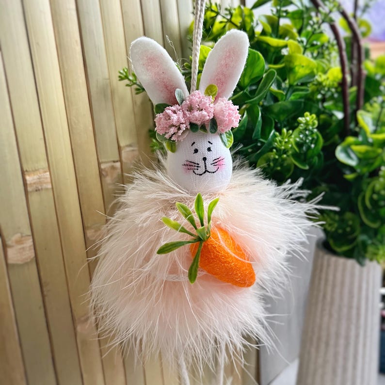 May include: A pink fluffy Easter bunny with a flower crown and a carrot in its hand. The bunny is hanging from a string.
