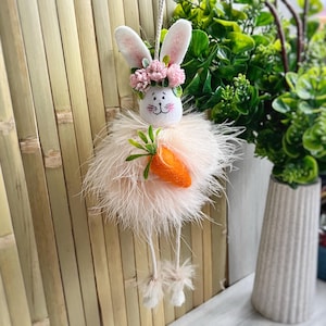 May include: A pink fluffy Easter bunny with white ears and a pink flower crown holds a carrot. The bunny has a white face with black eyes and a black nose. The bunny is hanging from a string.