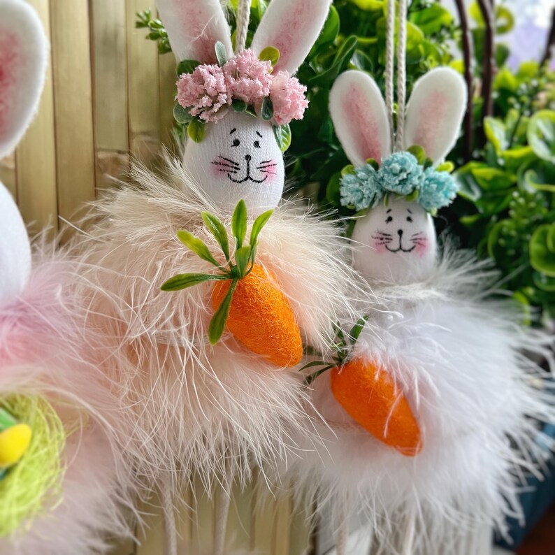 May include: Three white plush bunny ornaments with fluffy white feathers. Each bunny is wearing a flower crown and holding a carrot. The bunnies are hanging from a string.