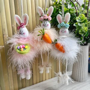 May include: Three plush Easter bunny ornaments with long legs and feathery skirts. One bunny holds a carrot, one holds a nest of colorful eggs, and one has a flower crown.