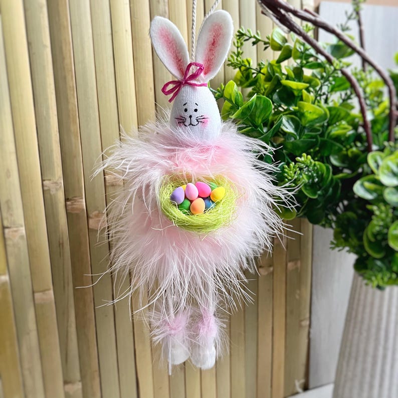 May include: A pink Easter bunny with long feathery legs holds a nest filled with colorful Easter eggs.