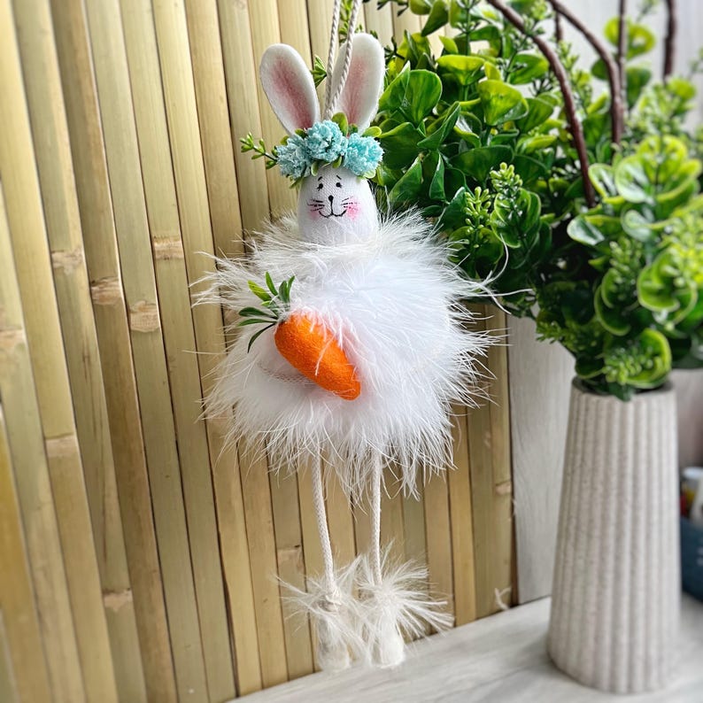 May include: White fluffy Easter bunny with long legs holding a carrot. The bunny is wearing a blue flower crown and has a painted face.