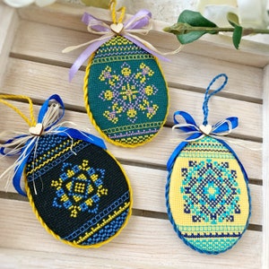 Peut inclure: Trois œufs de Pâques brodés avec des motifs géométriques différents. Les œufs sont décorés de fils jaunes, bleus, verts et noirs. Chaque œuf est orné d'un ruban bleu avec un petit charme en forme de cœur.