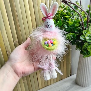 May include: Pink Easter bunny with white ears and a pink bow holding a green nest filled with colorful eggs.