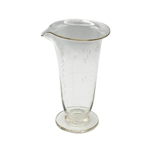 May include: A clear glass beaker with a flared top and a pouring spout. The beaker has measurement markings and sits on a circular base. The rim has a gold-colored trim.