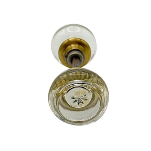 May include: A clear glass doorknob with a brass stem and a starburst design on the bottom knob. The top knob is round and clear. The doorknob is likely for a vintage or antique door.