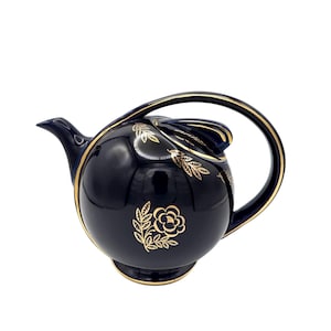 May include: A dark blue teapot with a gold floral design and trim. The teapot has a rounded shape with a curved handle and a spout. The gold floral design includes a rose and leaf pattern.