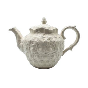 May include: A cream-colored ceramic teapot featuring a textured pineapple-inspired surface pattern. The teapot includes a matching lid with a small finial, a curved spout, and a decorative handle, all finished in a uniform glossy off-white glaze.