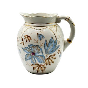 May include: A white ceramic pitcher featuring a scalloped rim with gold trim. The body is decorated with hand-painted blue flowers and brown foliage. A curved handle with gold accents extends from the side, and the base has a rounded shape.