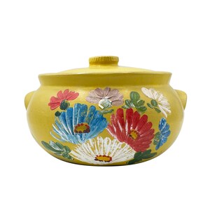 May include: A yellow ceramic casserole dish with a lid and two side handles. The exterior features a hand-painted floral design with red, blue, and white flowers and green leaves arranged in a cluster across the front of the rounded body.