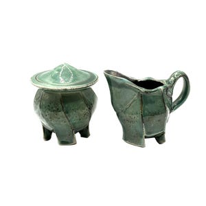 May include: A two-piece ceramic set in a teal green glaze. The set includes a lidded sugar bowl and a creamer with a handle. The sugar bowl has a rounded body and three feet. The creamer has a unique, angular design.