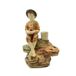 May include: A ceramic figurine of a person wearing a dark hat, tan shirt, and brown pants, sitting barefoot on a textured, multi-colored rock formation. The rock base features several circular openings and a small cylindrical pillar on the top surface.