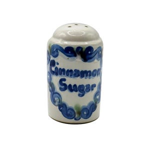 May include: A white ceramic shaker with a rounded top and perforated lid. The body features hand-painted blue scrollwork framing the text Cinnamon Sugar in blue script. The cylindrical container has a smooth, glossy finish and a flat base.