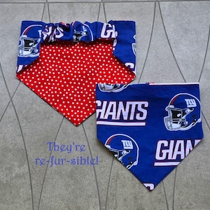 May include: Two reversible dog bandanas. One is blue with white football helmets and the word "GIANTS". The other side is red with white stars. The second bandana is blue with white football helmets and the word "GIANTS". The text "They're re-fur-sible!" is also visible.