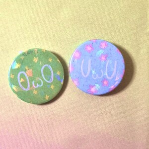 Op de afbeelding: Twee ronde pinnen met verschillende ontwerpen. De eerste pin is groen met gouden sterren en de tekst "OwO". De tweede pin is blauw met roze bloemen en de tekst "uwu".